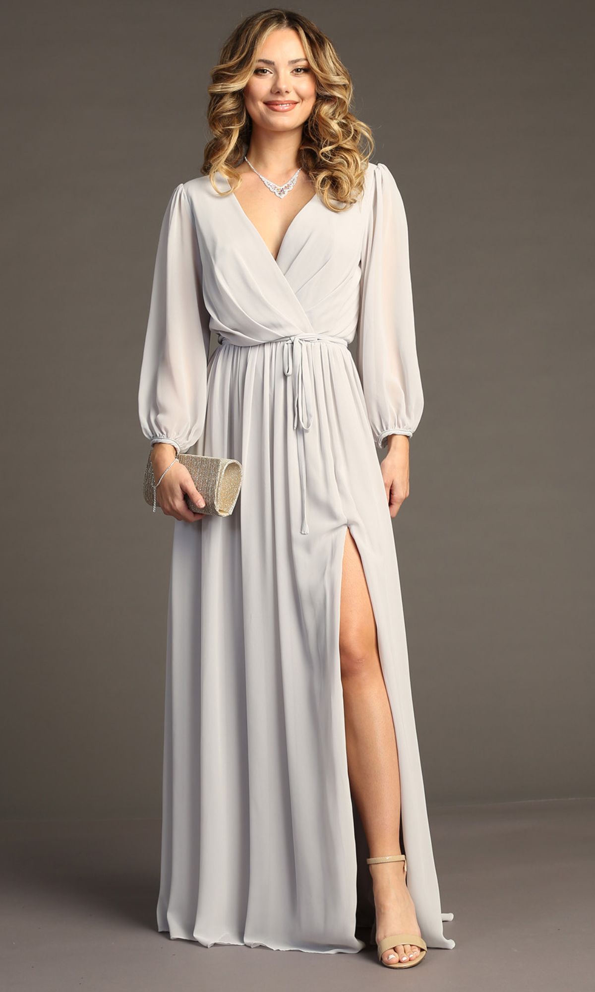A woman in the Chicas J012B Long-Sleeve Long Formal Dress—a light gray gown with a wrap design and thigh-high slit—poses against a plain background, styled with heels, a clutch, and a necklace.